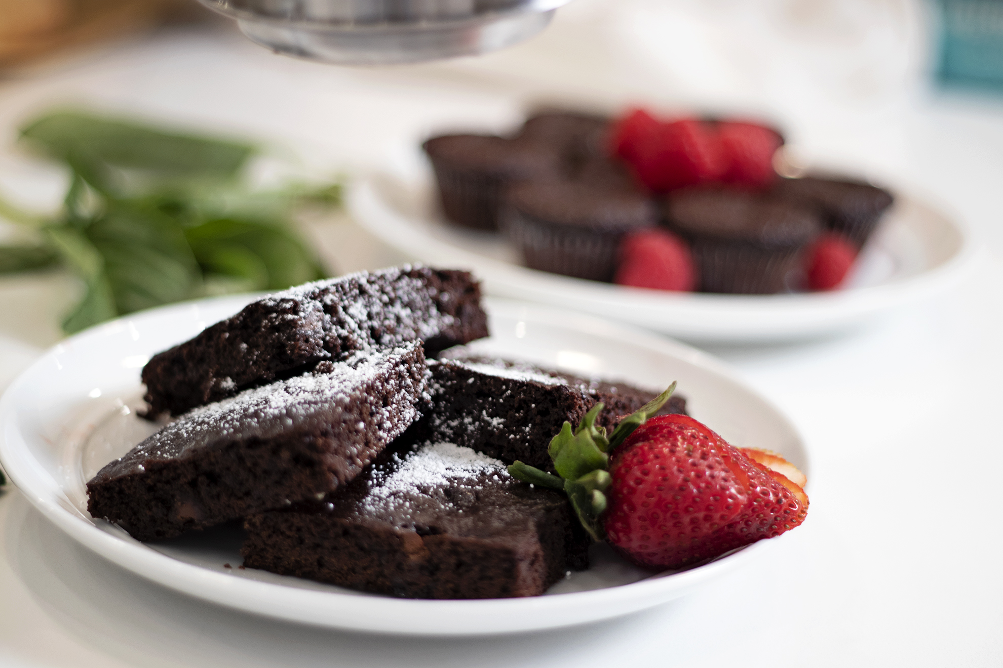 Photo of brownies on a plate with powdered sugar and a strawberry with chocolate muffins and rasperries in the background